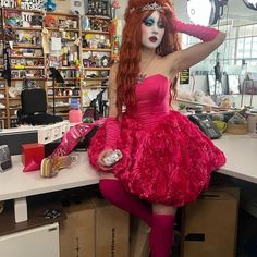 a woman with red hair and makeup is sitting on a counter in a store wearing a pink dress