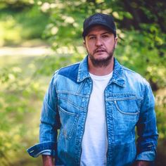 a man wearing a denim jacket and hat standing in front of some trees with his hands in his pockets