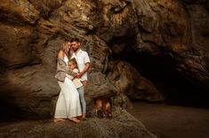 a man and woman standing next to a brown dog on a rock covered beach with a cave in the background