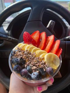 a person holding up a bowl of fruit and granola in their hand while driving