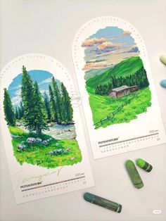 two calendars with colored pencils next to them on a white surface, one has an image of a cabin and the other shows mountains