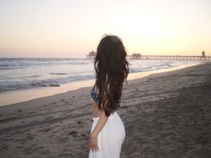 a woman standing on top of a sandy beach next to the ocean with her hair blowing in the wind