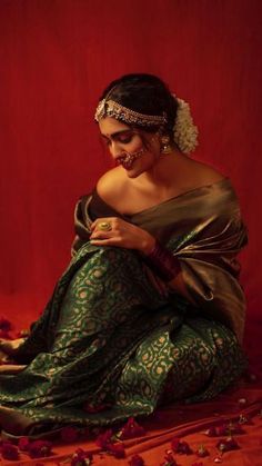 a woman in a green and gold sari sitting on a red background with rose petals