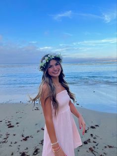 a beautiful young woman standing on top of a beach next to the ocean wearing a flower crown