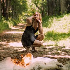 a woman kneeling down next to a fire in the middle of a forest with her hand on her face