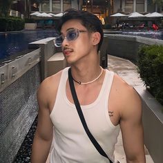 a young man wearing sunglasses and a white tank top standing in front of a pool