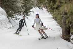 two people skiing down a snow covered slope in the woods with trees on either side