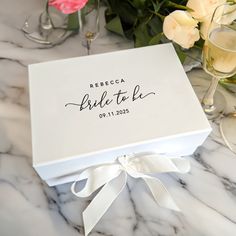 a white box with the words, bridal to be on it sitting next to some wine glasses and flowers