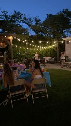 two women sitting at a table in the grass with lights strung over them and people standing around