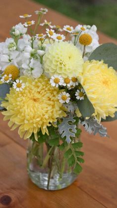 a glass vase filled with yellow and white flowers on top of a wooden table next to a green leaf