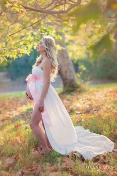 a pregnant woman in a white dress poses for the camera while standing under a tree