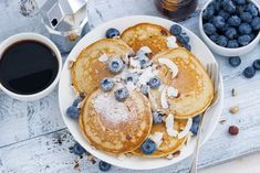 pancakes with blueberries and powdered sugar are on a white plate next to cups of coffee