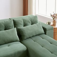 a green couch sitting in front of a window next to a wooden table with flowers on it