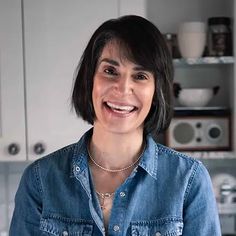 a smiling woman in a denim shirt stands in front of a kitchen with white cabinets