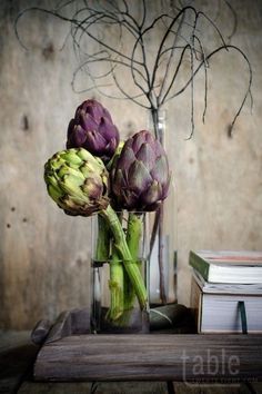 three artichokes in a glass vase on a table next to two books