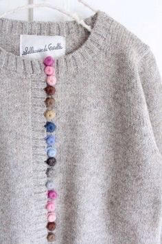 a gray sweater with multicolored beads hanging on a hanger next to a white wall