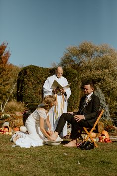 a bride and groom sitting on the grass at their outdoor wedding ceremony with priest in background