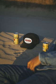 two cans of beer and a hat sit on the sand next to a man's feet