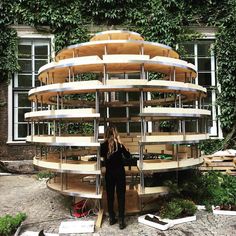 a woman standing in front of a circular wooden structure with stairs on the side and plants growing around it