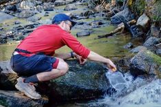 a man in red shirt and blue hat kneeling on rocks next to stream with water