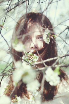 a woman with long brown hair and blue eyes is looking at the camera while she has flowers in her mouth