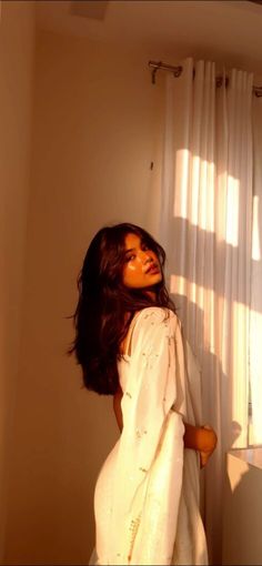 a woman standing in front of a window next to a white curtain with the sun shining through it