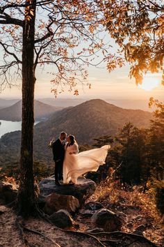 a bride and groom standing on top of a mountain with the sun setting behind them