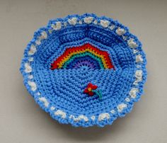 a blue crocheted bowl with a rainbow in the center on a white surface