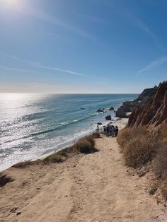 a group of people walking down a sandy path next to the ocean on a sunny day