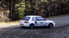 a white car driving down a dirt road in front of some pine trees and bushes