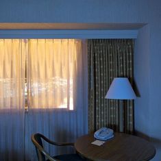 a hotel room with a table, chair and lamp next to a window that has curtains on it