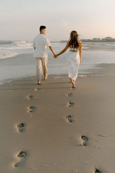 a man and woman holding hands walking on the beach with footprints in the sand as the sun sets