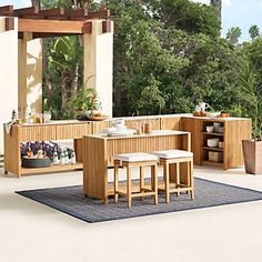 an outdoor kitchen with wooden furniture and potted plants on the table, in front of trees