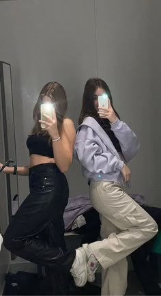 two women are taking pictures in the mirror with their cell phones while they both wear pants and sneakers