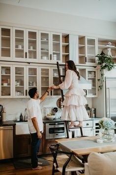 a man and woman standing on ladders in a kitchen next to a stove top oven