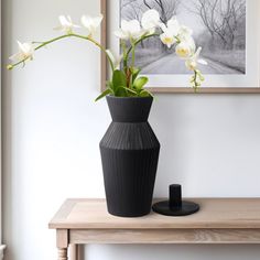 a black vase with white flowers sitting on a wooden table next to a framed photograph