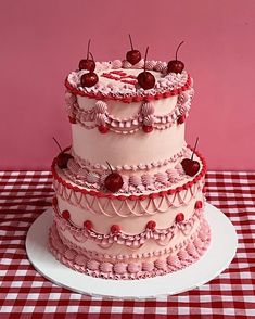 a three tiered cake with cherries on top is sitting on a checkered tablecloth