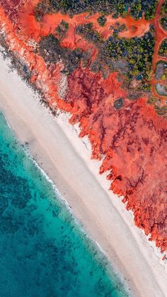 an aerial view of the beach and ocean with red sand, blue water and trees