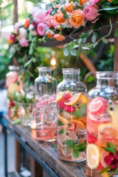 several vases filled with different types of fruit and flowers on a wooden table surrounded by greenery