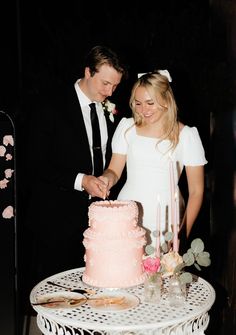 a man and woman cutting into a pink cake on top of a white table next to each other