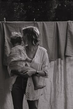 a black and white photo of a woman holding a baby in her arms while standing next to a clothes line