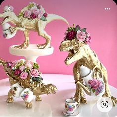 two gold dinosaur figurines sitting on top of a white cake plate with pink flowers