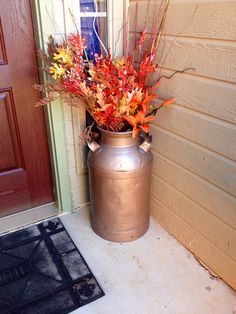 a metal container with flowers in it sitting next to a door mat on the side of a house