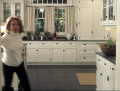 a woman standing in the middle of a kitchen with white cabinets and black counter tops