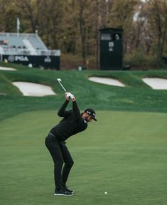 a man in black playing golf on a green field with trees and grass behind him