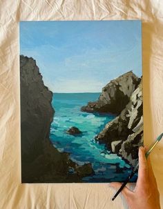 a person holding a paintbrush in their hand near a painting of the ocean and rocks