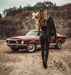 a woman walking down a dirt road next to a red car