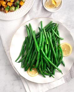 a white plate topped with green beans next to a bowl of lemon wedged potatoes