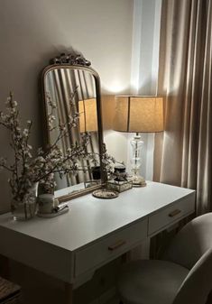 a white desk with a mirror, lamp and flowers on it in front of a window