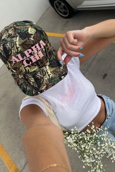 a woman in shorts and a camo hat holds flowers up to the camera with her right hand